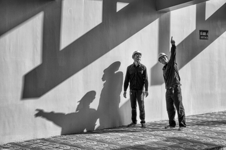 Two engineers with helmets cast shadows while inspecting a construction site with dynamic lighting.