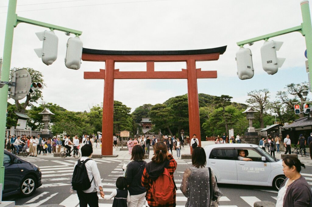 People crossing at a busy intersection under a large red torii gate in Kamakura, Japan.